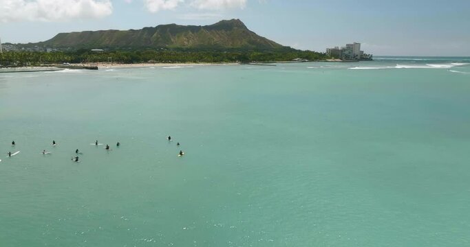 Flying backwards above the ocean surface to reveal surfers on the surfboards and panorama of Damon Head volcanic peak in Honolulu, Hawaii 