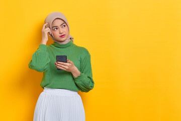 Pensive beautiful Asian woman in green sweater holding mobile phone looks seriously thinking about...