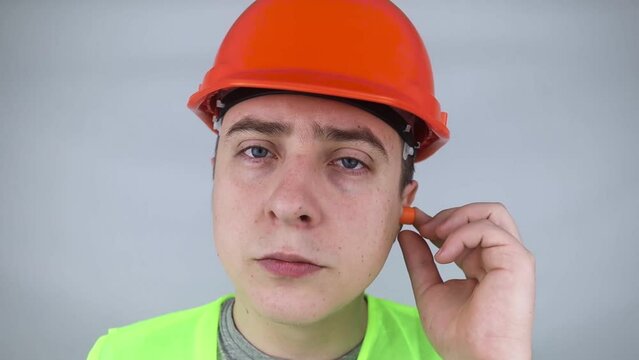 A worker in a hard hat and green vest puts on ear plugs. Industrial safety. Industrial noise protection. Compliance with the rules of conduct at loud work. Orange ear plugs