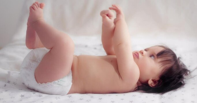 Cute funny baby girl with dark hair is lying in a diaper on her back, on the bed in the nursery moving her legs and arms. The concept of love, parenthood, childhood, life, motherhood