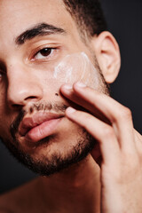 Close-up of bearded young man applying face cream on his face and looking at camera - obrazy, fototapety, plakaty