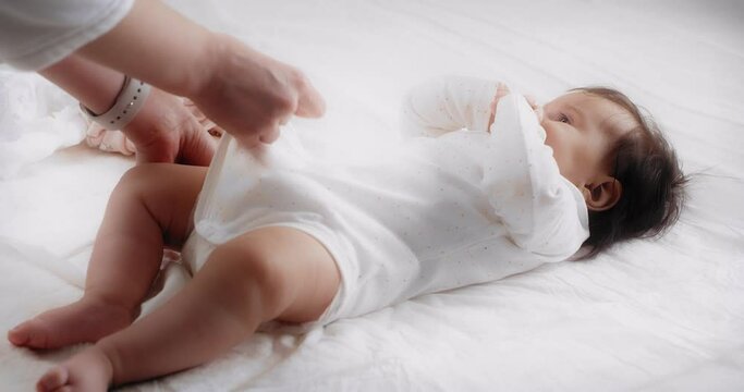 Mother's caring hands put a white bodysuit on her little daughter. Baby girl is lying on her back on a white bed in the nursery. Mom gently strokes the baby