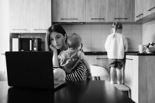 a young mother with many children working on a laptop sitting in the kitchen with a baby in her arms while the eldest daughter helps with cooking and cleaning in the kitchen