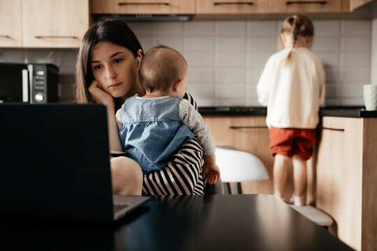 a young mother with many children working on a laptop sitting in the kitchen with a baby in her arms while the eldest daughter helps with cooking and cleaning in the kitchen