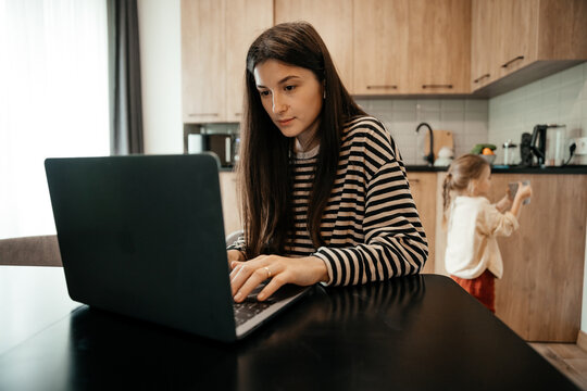 Young female freelancer mother dressed in home clothes working on laptop sitting at the table. The child in the background is playing in the kitchen