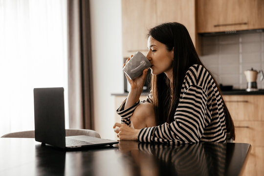 Young female freelancer dressed in home clothes working on laptop sitting at the table in the kitchen. Stylish girl on workplace at the desk. Distance learning online education