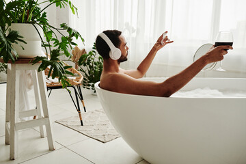Young man relaxing in bathtub with glass of red wine and enjoying music in headphones during spa...