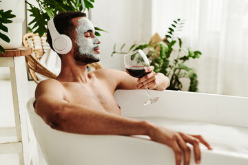 Young man enjoying relaxed time in bathroom, he listening to music in wireless headphones and enjoying the glass of red wine