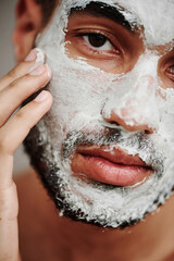 Close-up of handsome young man applying cosmetic mask on his face and looking at camera