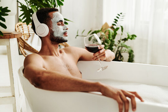Young man enjoying relaxed time in bathroom, he listening to music in wireless headphones and enjoying the glass of red wine