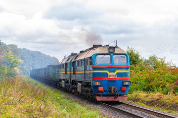 A powerful blue diesel locomotive pulls a long freight train loaded with coal along the railway tracks. Autumn photo. International Freight rail transport