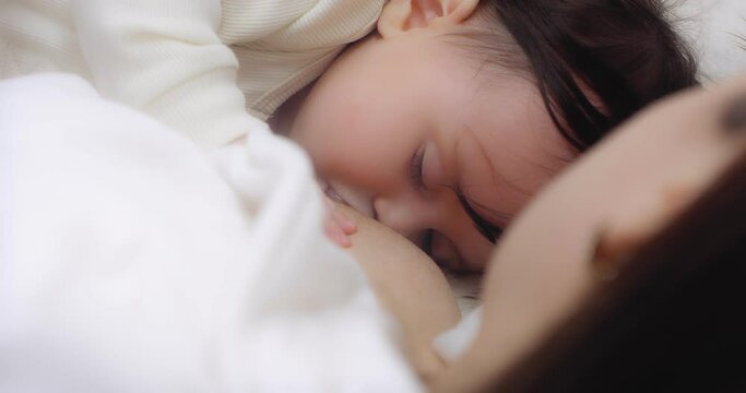 Mother in a white coat is breastfeeding her sleeping little daughter lying on the bed. Baby sucks milk from mother's breast. Concept of love, parenthood, childhood, life, motherhood