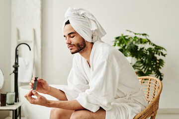 Young metrosexual man in bathrobe sitting on chair in bathroom and taking care of his hands