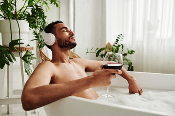 Young mixed race man listening to music in wireless headphones and drinking wine while relaxing in bath