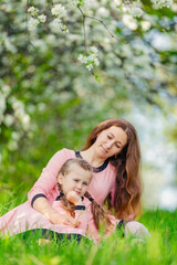 Fototapeta premium mother and daughter sit in the green grass against the backdrop of blooming apple trees