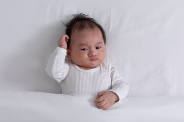 Top view happy newborn baby lying and scratching head thinking something on a white bed and blanket...