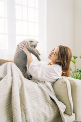 Young girl enjoy playing with  cat on sofa, holding happy soft cuddly white cat at home warm and cozy minimal atmosphere