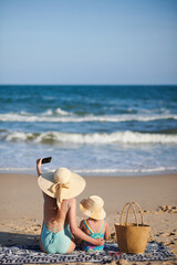 Mother and daughter in straw hats sitting on beach and taking selfie on smartphone, view from the back