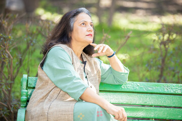 Thinking Indian asian senior woman sitting on bench at park, old retired female.copy space....