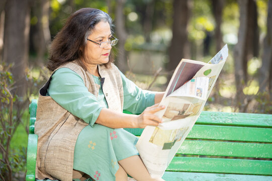 Indian or asian senior woman wearing eyeglasses reading newspaper while sitting on bench at park, Old Grey hair female relaxing at outdoor garden.