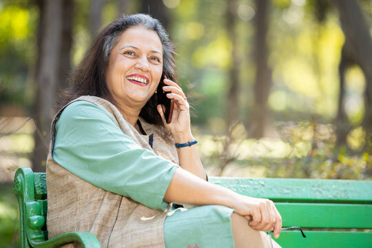 Happy Indian Senior Woman Talking On Mobile Phone While Sitting On Bench At Park Outdoor, Mature Old People Using Technology.