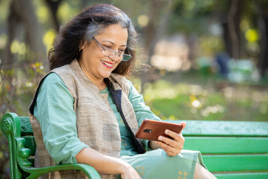 Happy Indian senior woman wearing eye glasses watching video or social media on smart phone while sitting on bench at park outdoor, Mature old people using technology.