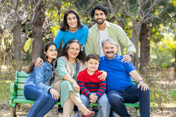 Portrait of Happy indian family sitting together on a bench in a park. Asian senior and young couple with their kids wearing casual cloths smiling, enjoying picnic holiday outdoor. selective focus
