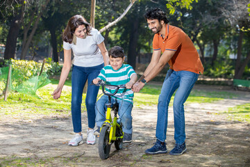 Indian mother and father helping son learn to ride a bicycle in the park outdoor. kid having fun...
