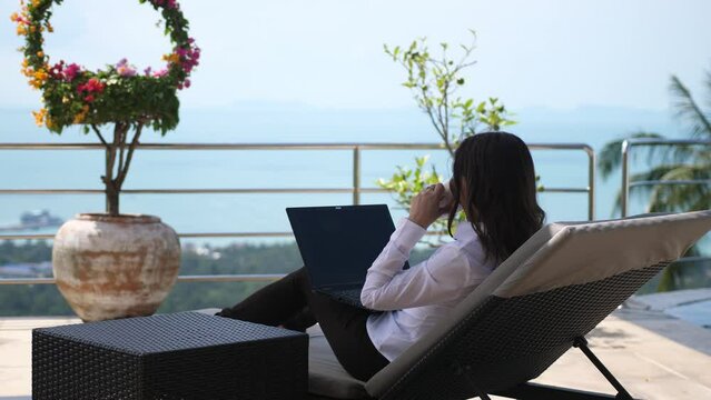 Woman lying on a sunbed on the terrace of a villa working with a laptop overlooking the sea view. freelance work