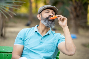 Indian senior man eating licking ice lolly or ice cream in a park outdoor, mature bead male enjoy...