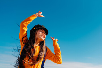 cheerful asian girl in yellow sweater and backpack making peace symbol with her hands while dancing...