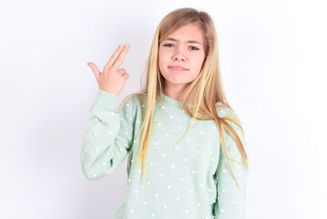 Unhappy little caucasian kid girl wearing fashion sweater over blue background makes suicide gesture and imitates gun with hand, curves lips, keeps two fingers on temple