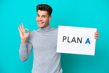 Young handsome caucasian man isolated on blue bakcground holding a placard with the message PLAN A with ok sign