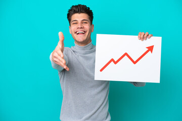 Young handsome caucasian man isolated on blue bakcground holding a sign with a growing statistics arrow symbol making a deal