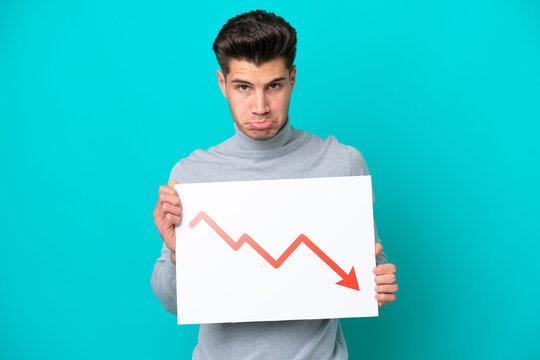 Young handsome caucasian man isolated on blue bakcground holding a sign with a decreasing statistics arrow symbol with sad expression