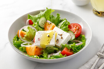 Pouring olive oil on greek salad with green leaves, tomatoes, cucumber, feta cheese. White bowl, marble