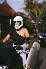 Fototapeta na wymiar A young woman in a motorcycle full face helmet rides a motobike. Surfer on a motorcycle, surfboard in racks. Safe driving in Asia.