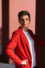 A stylish young business woman with short red hair in a red jacket, white T-shirt and black trousers stands in the courtyard of the theater building with red columns. Interesting shadows in red tones