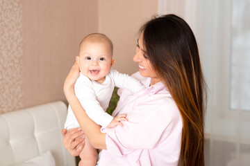 A portrait of a beautiful young mother and her sweet baby in light clothes in their bedroom at home. Lifesthal. Motherhood. Happy family