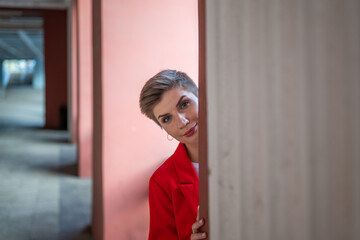 A stylish model girl with short brown hair in a red jacket and a white T-shirt, hiding behind a concrete square column, peeking out from behind it (peeping or spying)