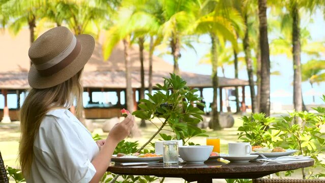 Young woman in hat eat watermelon in outdoor resort restaurant. Girl have healthy breakfast with fresh fruits on terrace of cafe of luxury hotel on tropical island. Vacations or holidays concept.