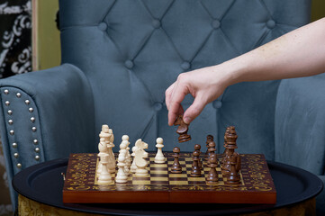 Close-up of a man's hand holding a chess figure under the comfortable vintage blue coloured armchair over full of other different coloured figures standing on the wooden chessboard playing game.