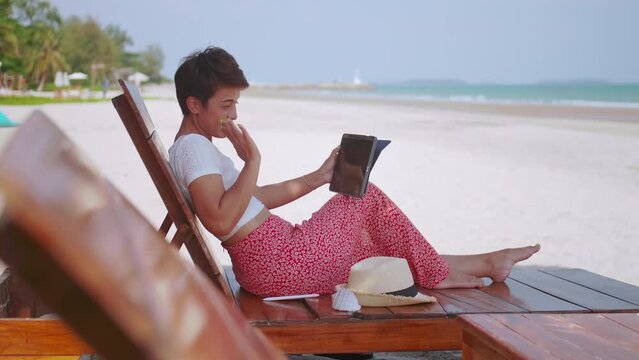 Asian woman chatting with family on video call while relaxing on beach.