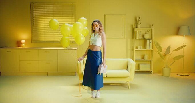 Full length of a trendy and carefree woman posing with a gift box and balloons in the studio with monochrome yellow interior. A cheerful female model holds balloons and a birthday present