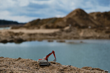 Small plastic toy excavator with bucket working on sand extraction at quarry. Pond in background. Children's toy model of tractor extracts sand. Construction and mining industry. Creative banner.