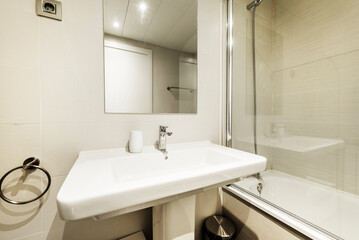 Fototapeta na wymiar Bathroom with white porcelain sink on porcelain pedestal, frameless mirror, glass-enclosed tub, and light tile
