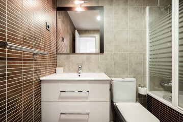 Fototapeta na wymiar Bathroom with white resin sink on wooden cabinet with drawers, mirror on the wall, shower cabin and brown tiles