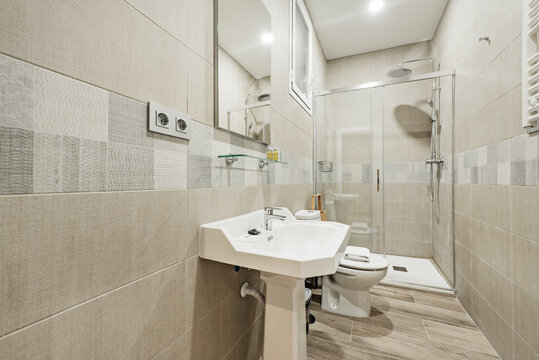 Bathroom with white porcelain sink on porcelain pedestal, frameless mirror, glass-enclosed shower stall, wide light tiled valance