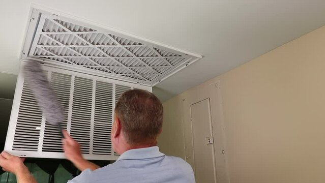 Mature male dusting the inside and outside of his air intake grid in a home furnace ceiling. Entryway ceiling air intake vent to furnace large grid being dusted inside and out in a home
