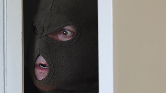A portrait of a criminal in a balaclava who opens the door and shocked looks into the camera